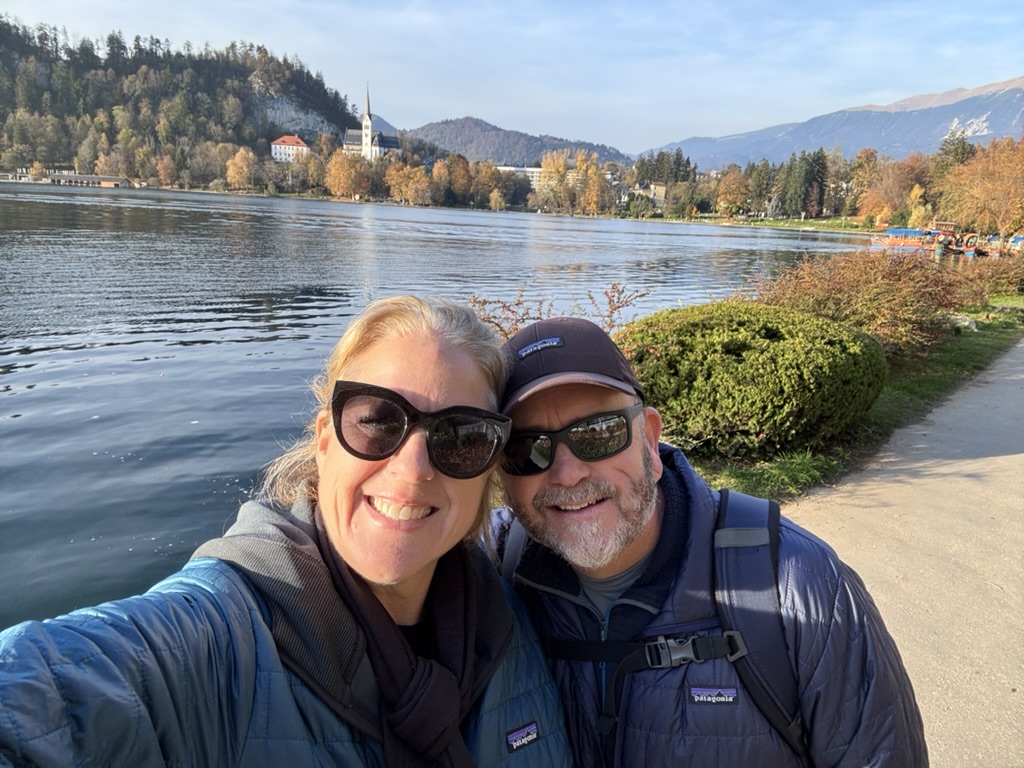 Susan Green and her husband at Lake Bled, Slovenia, with the church and autumn trees across the water