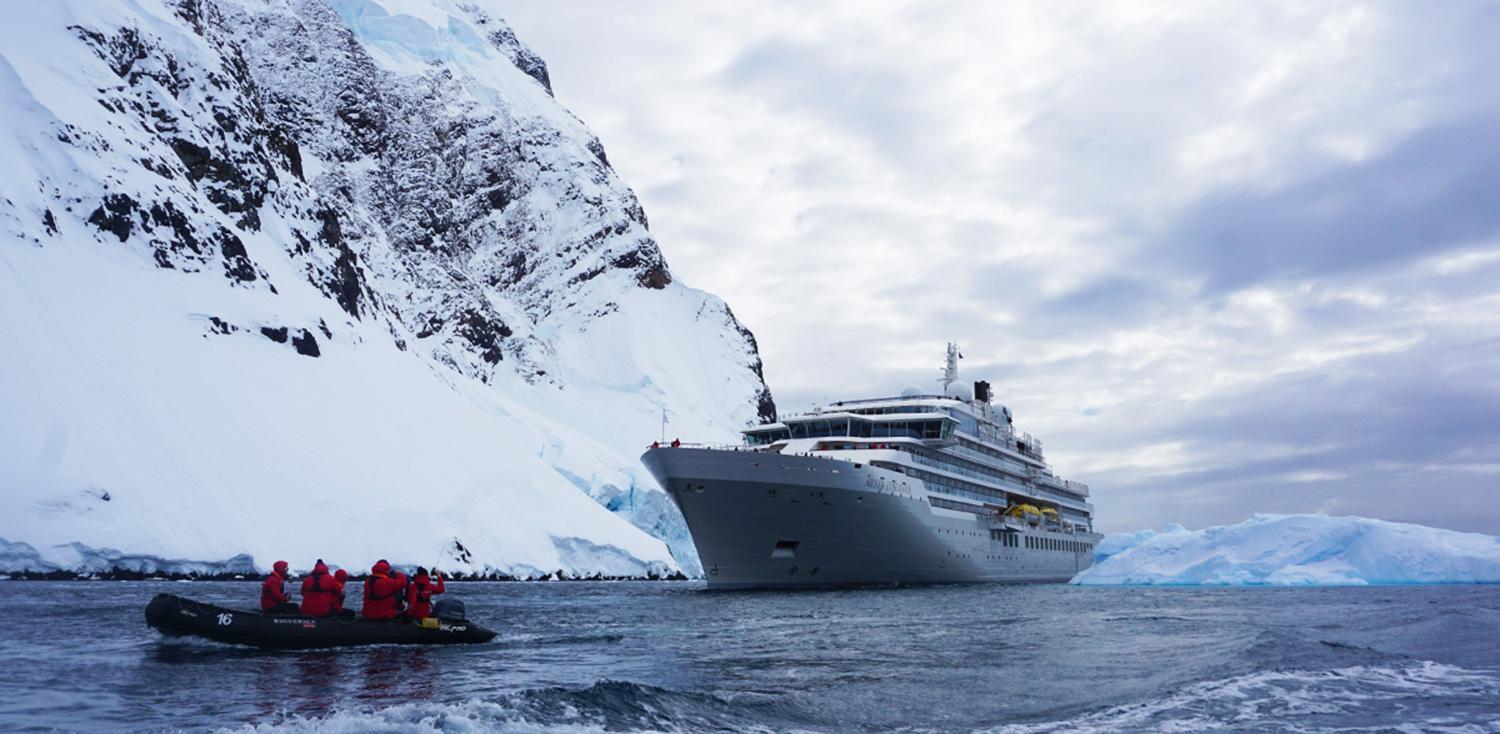 Silversea expedition ship and a Zodiac of guests in red parkas below a snowy Antarctic cliff
