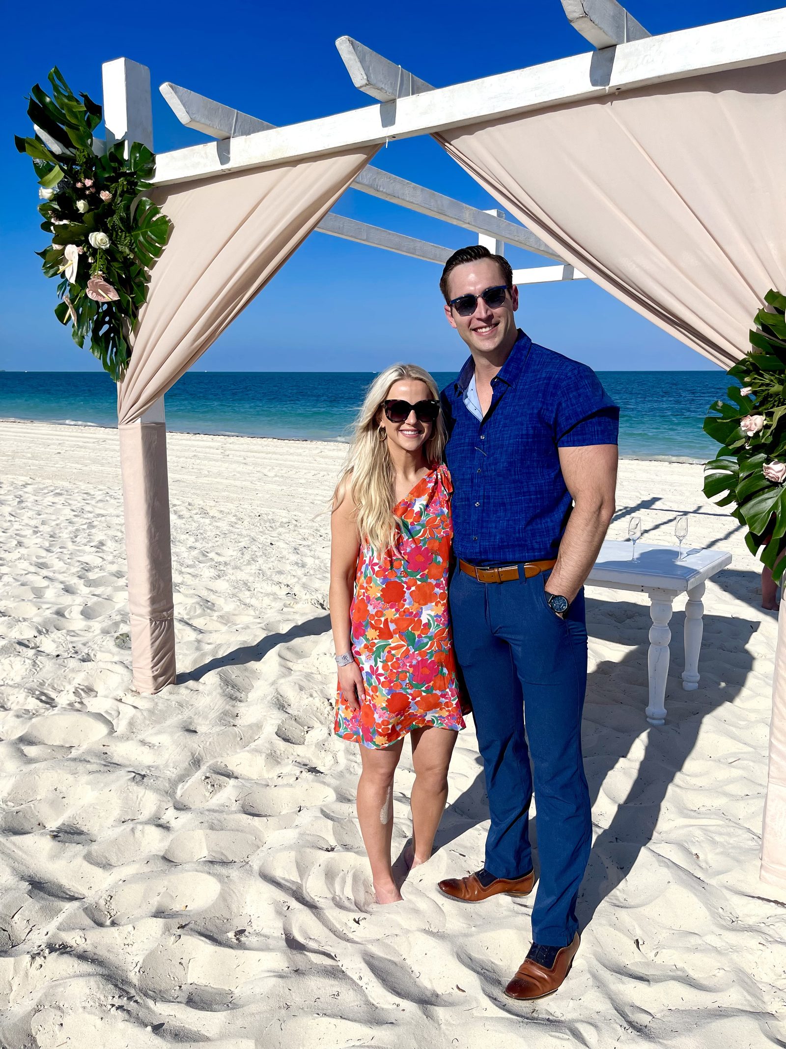 Haley and her husband Ryan smiling at a wedding arch on a white-sand beach