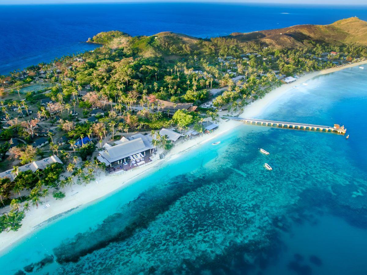 Aerial view of a Mamanuca Island in Fiji, with a white-sand beach, palm trees and a long pier over turquoise water